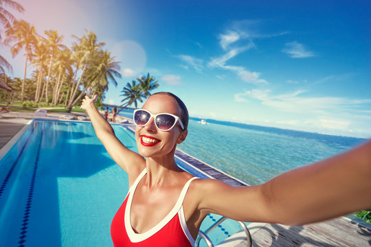 Vacation and technology. Colorful portrait of pretty young woman taking selfie portrait near swimming pool on tropical beach.