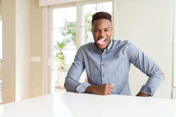 Fototapeta premium Handsome african american man on white table sticking tongue out happy with funny expression. Emotion concept.