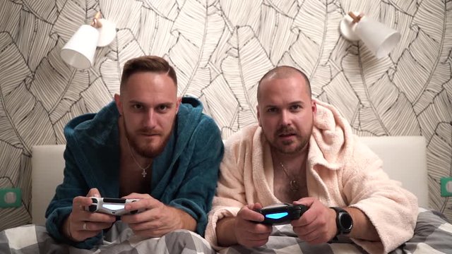 Two guys play computer games on the bed in bathrobes and win. Slow motion footage 2 bearded men play video game