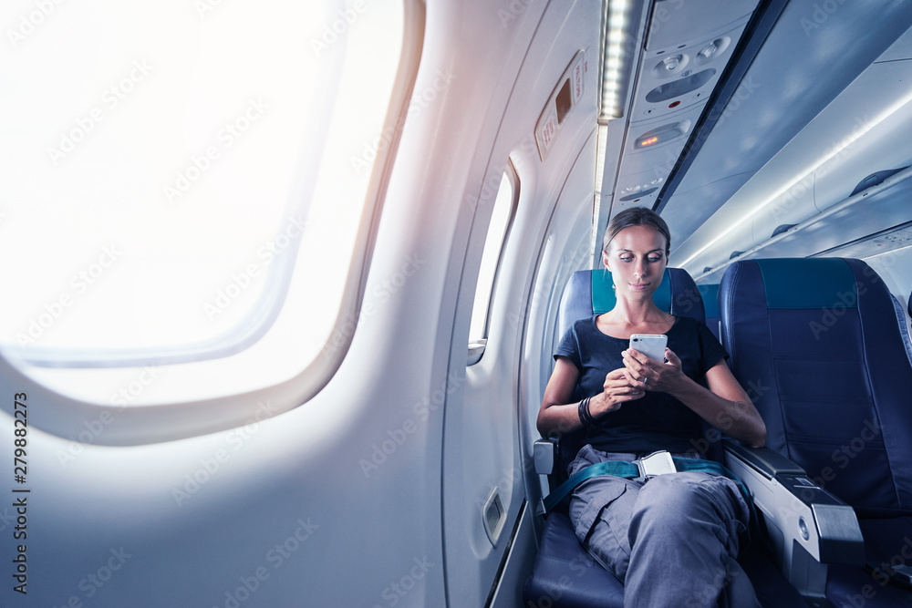 Wall mural travel and technology. young woman in plane using smartphone while sitting in airplane seat.