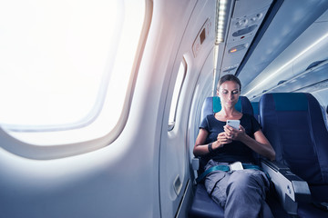 Travel and technology. Young woman in plane using smartphone while sitting in airplane seat.