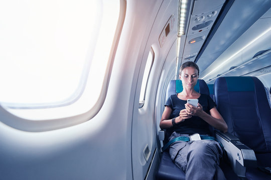 Travel And Technology. Young Woman In Plane Using Smartphone While Sitting In Airplane Seat.