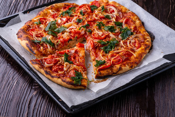 Freshly cooked hot pizza and slice with cheese ham meat tomato onion parsley homemade on parchment paper in baking sheet tray recipe on dark wooden table