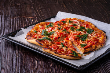 Freshly cooked hot pizza and slice with cheese ham meat tomato onion parsley homemade on parchment paper in baking sheet tray recipe on dark wooden table