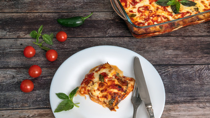 Eggplant parmegiano, a piece on a plate with a knife and fork, next to the tray with eggplants, cherry tomatoes