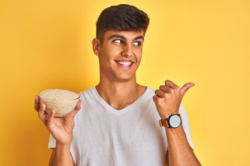 Young indian man holding bowl with rice standing over isolated yellow background pointing and showing with thumb up to the side with happy face smiling