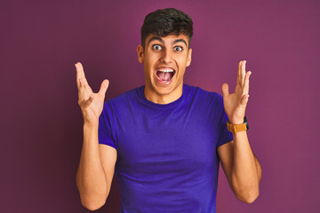 Young indian man wearing t-shirt standing over isolated purple background celebrating crazy and amazed for success with arms raised and open eyes screaming excited. Winner concept