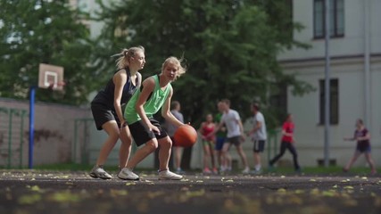 Tracking shot of two college girls playing basketball on outdoor court in slow motion. Female friends practicing defender and forward roles, group of students exercising in background - Powered by Adobe