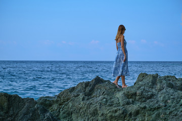 Naklejka premium A beautiful young girl in a blue dress is standing on a rock and looking at the sea.