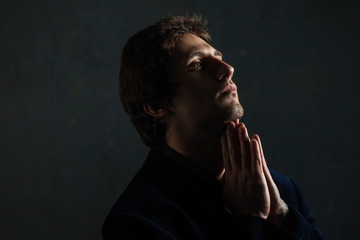 Portrait of a young pensive man on a dark background, looking to the side. Hands in prayer gesture.