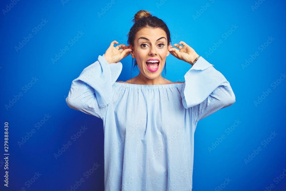 Canvas Prints Young beautiful woman wearing bun hairstyle over blue isolated background Smiling pulling ears with fingers, funny gesture. Audition problem