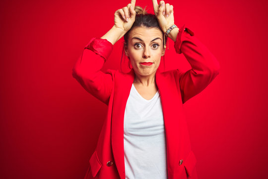 Young beautiful business woman standing over red isolated background doing funny gesture with finger over head as bull horns