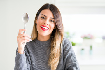 Young beautiful woman holding silver spoon at home with a happy face standing and smiling with a confident smile showing teeth
