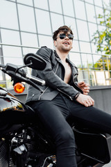 motorcyclist in sunglasses and black leather jacket sitting on motorcycle, smiling and looking away