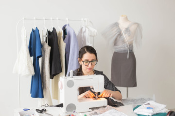 Dressmaker, tailor and fashion concept - Smiling female fashion designer using sewing machine and sitting behind her desk