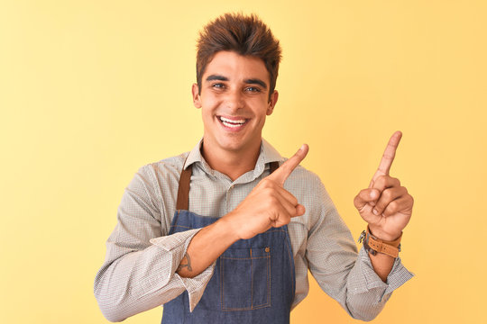Young handsome employee man wearing apron standing over isolated yellow background smiling and looking at the camera pointing with two hands and fingers to the side.