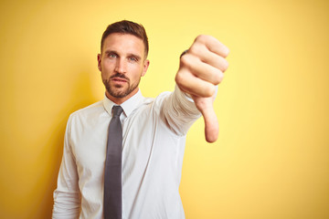 Young handsome business man wearing elegant white shirt over yellow isolated background looking unhappy and angry showing rejection and negative with thumbs down gesture. Bad expression.