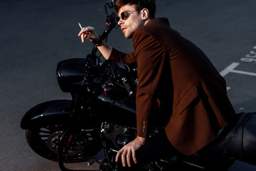 young man in brown jacket sitting on black motorcycle and smoking cigarette