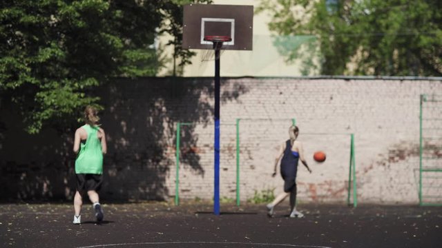 Two college girls practicing basketball shots on outdoor urban court. Back view of young female basketball player shooting hoop and scoring goal, her friend clapping hands