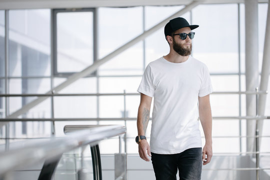 Hipster handsome male model with beard wearing white blank t-shirt and a baseball cap with space for your logo or design in casual urban style