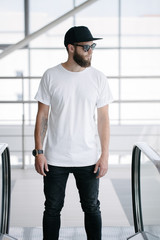Fototapeta na wymiar Hipster handsome male model with beard wearing white blank t-shirt and a baseball cap with space for your logo or design in casual urban style