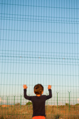one helpless little child refugee standing near border holding fencing with razor wire with copy...