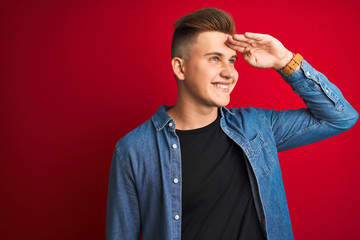 Young handsome man wearing denim shirt standing over isolated red background very happy and smiling looking far away with hand over head. Searching concept.