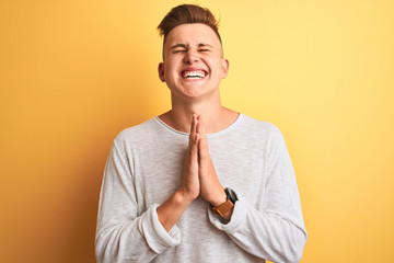 Young handsome man wearing white casual t-shirt standing over isolated yellow background begging...