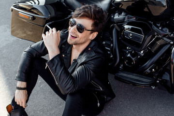 Fototapeta na wymiar young man in leather jacket sitting on ground near black motorcycle, smiling and smoking cigarette