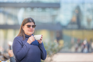 Lady is at a city cafe outdoors and drinks a cup of coffee.