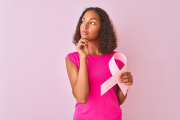 Young brazilian woman holding cancer ribbon standing over isolated pink background serious face thinking about question, very confused idea