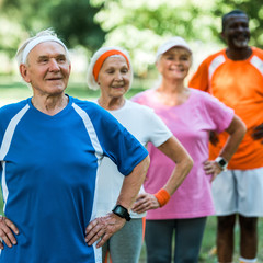 selective focus of cheerful retired man standing with hands on hips with multicultural pensioners in sportswear