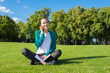 A smiling girl is sitting on the grass with a book in her hands, on a bright sunny day.