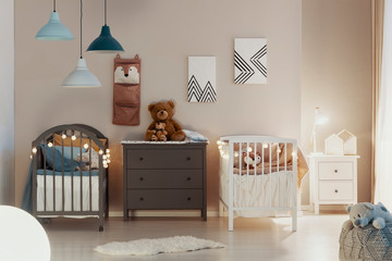 Real photo of a pastel bedroom interior for siblings with wooden cribs, beige walls, and cute teddy...