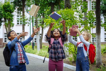 Back to school. Three Friends With Backpacks huging and laughing in front of school. Mixed Racial Group of School Kids having fun throwing up books in the schoolyard.