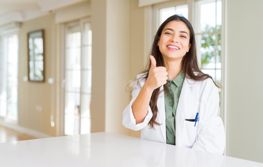 Young woman wearing medical coat at the clinic as therapist or doctor doing happy thumbs up gesture...