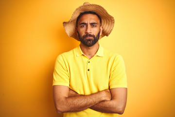 Young indian man on vacation wearing summer hat standing over isolated yellow background skeptic and nervous, disapproving expression on face with crossed arms. Negative person.