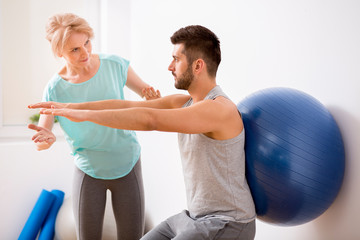 Young man with back injury exercising with blue gymnastic ball during appointment with female...