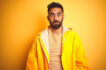 Young indian man wearing raincoat standing over isolated yellow background puffing cheeks with funny face. Mouth inflated with air, crazy expression.