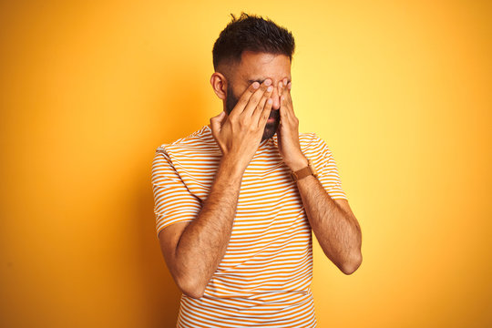 Young indian man wearing t-shirt standing over isolated yellow background rubbing eyes for fatigue and headache, sleepy and tired expression. Vision problem
