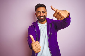 Young indian man wearing purple sweatshirt standing over isolated pink background approving doing positive gesture with hand, thumbs up smiling and happy for success. Winner gesture.