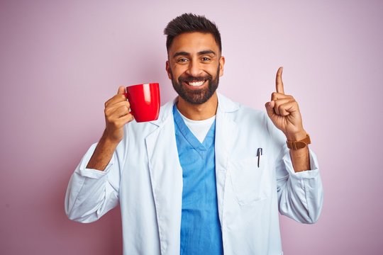 Young indian doctor man drinking cup of coffee standing over isolated pink background surprised with an idea or question pointing finger with happy face, number one
