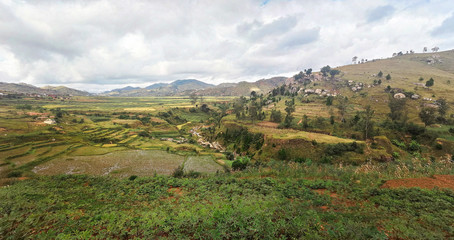 Fototapeta na wymiar Typical Madagascar landscape in region near Ambositra small hills covered with green grass and bushes, red clay houses and wet rice fields near.