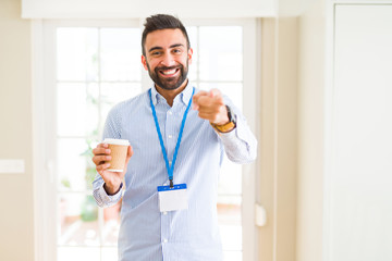 Handsome hispanic man wearing id card and drinking a cup of coffee pointing with finger to the camera and to you, hand sign, positive and confident gesture from the front