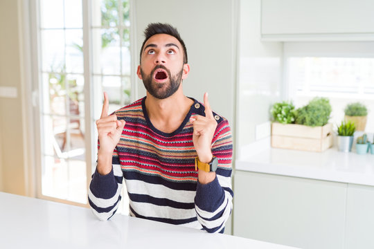 Handsome hispanic man wearing stripes sweater at home amazed and surprised looking up and pointing with fingers and raised arms.