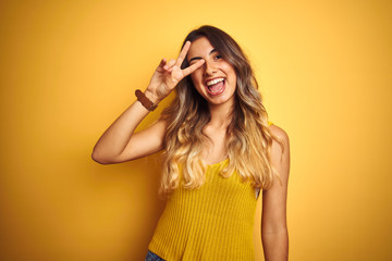 Young beautiful woman wearing t-shirt over yellow isolated background Doing peace symbol with fingers over face, smiling cheerful showing victory