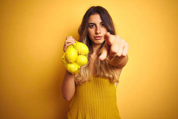 Young beautiful woman holding net of lemons over yellow isolated background pointing with finger to...