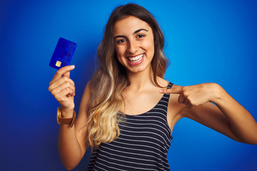 Young beautiful woman holding credit card over blue isolated background with surprise face pointing finger to himself