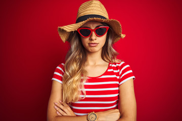Young beautiful woman wearing sunglasses and summer hat over red isolated background skeptic and nervous, disapproving expression on face with crossed arms. Negative person.