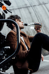 selective focus of man in sunglasses and brown jacket drinking alcohol from bottle while sitting on motorcycle